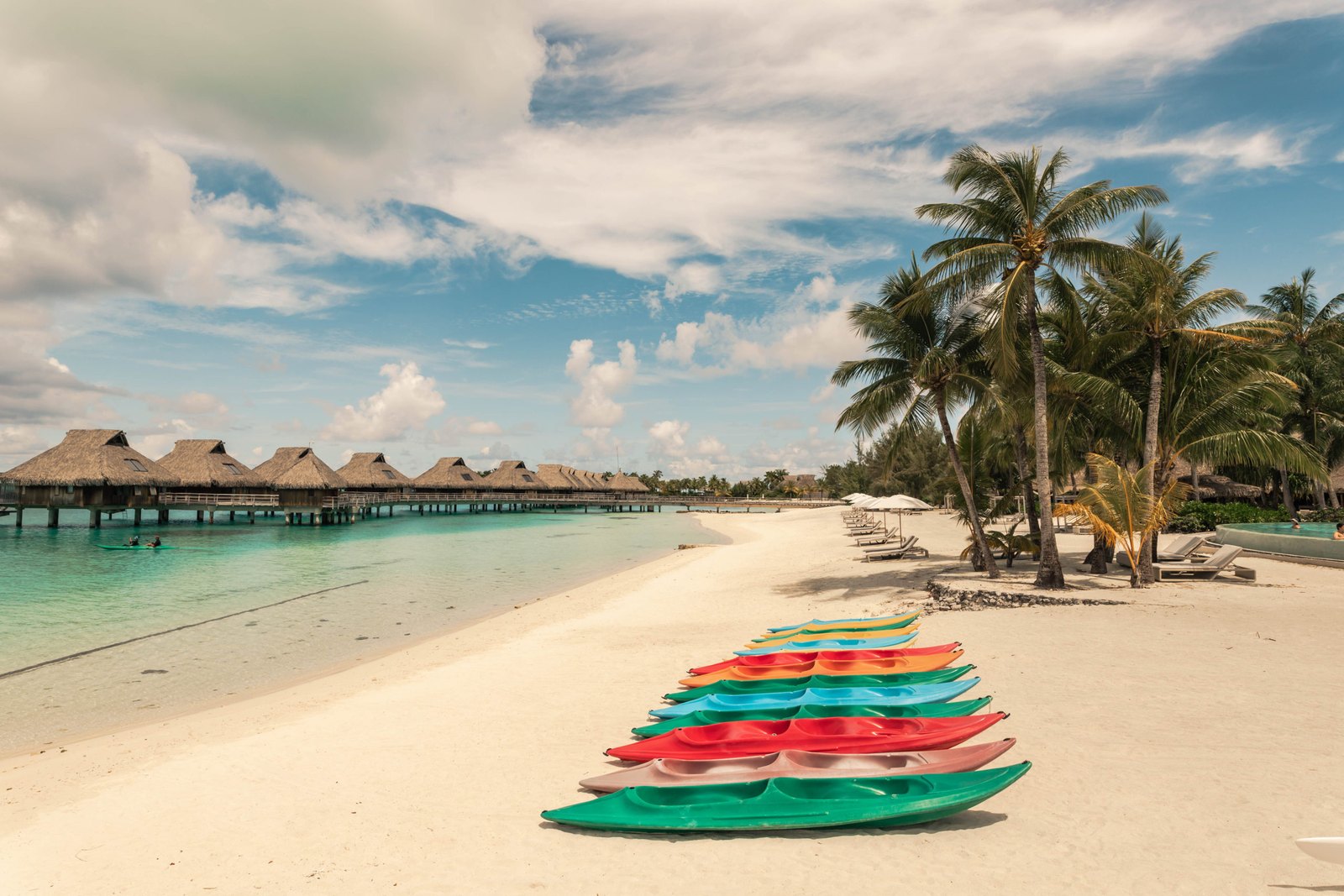 A row of colorful kayaks lined up on a white-sand beach, with palm trees and overwater bungalows stretching along the lagoon.