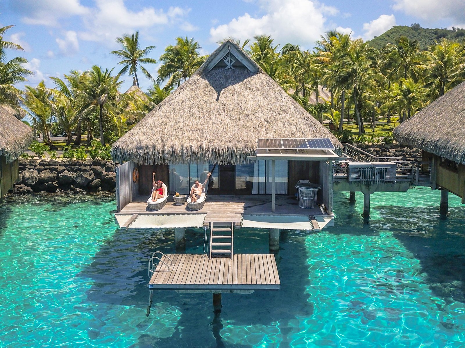 A thatched-roof overwater villa with two people relaxing on lounge chairs on the deck, a ladder to the water and a wooden platform over clear turquoise lagoon at the Conrad Bora Bora Nui.