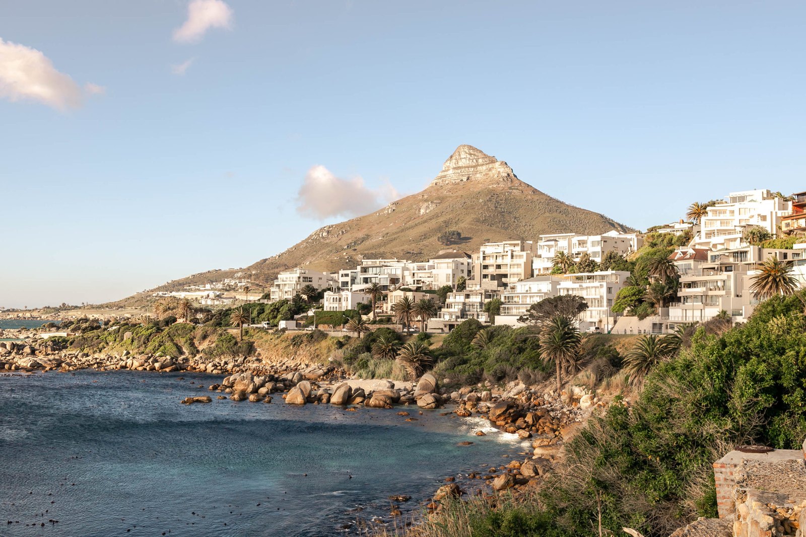 Camps Bay's white houses and rocky shoreline below Lion's Head under a blue sky.