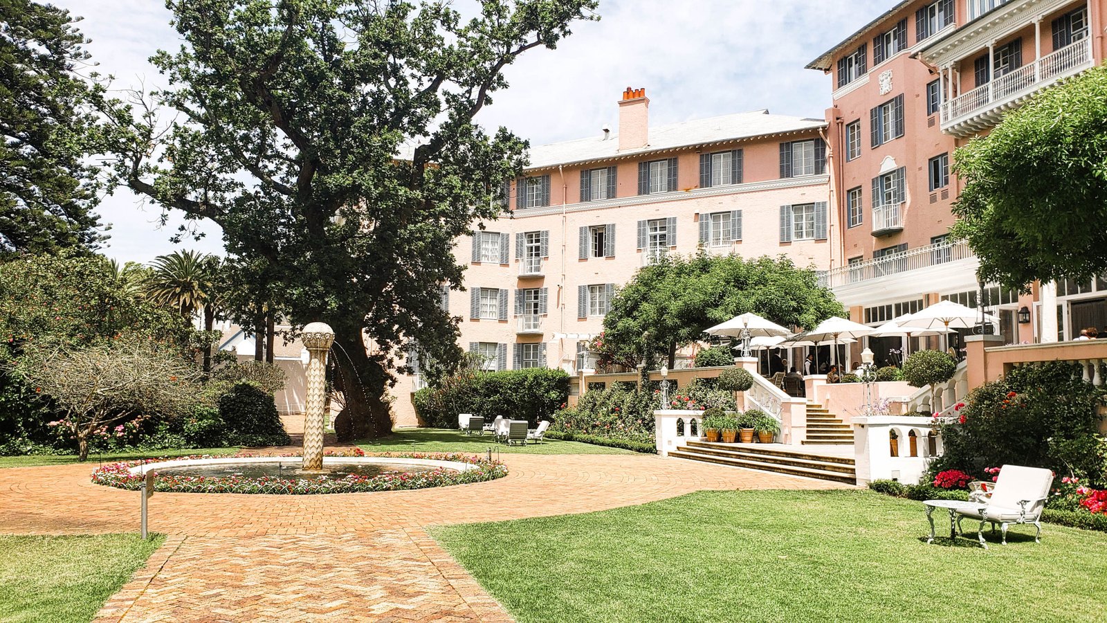 The pink Belmond Mount Nelson hotel behind a lawn and old tree in Cape Town.