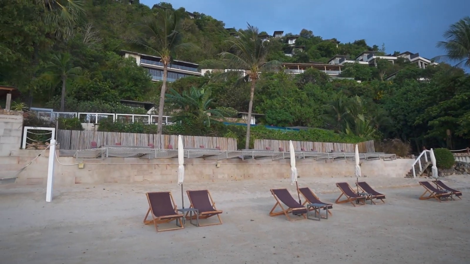 A sandy beach with wooden loungers and closed umbrellas, and villas stepping up a green hillside behind.