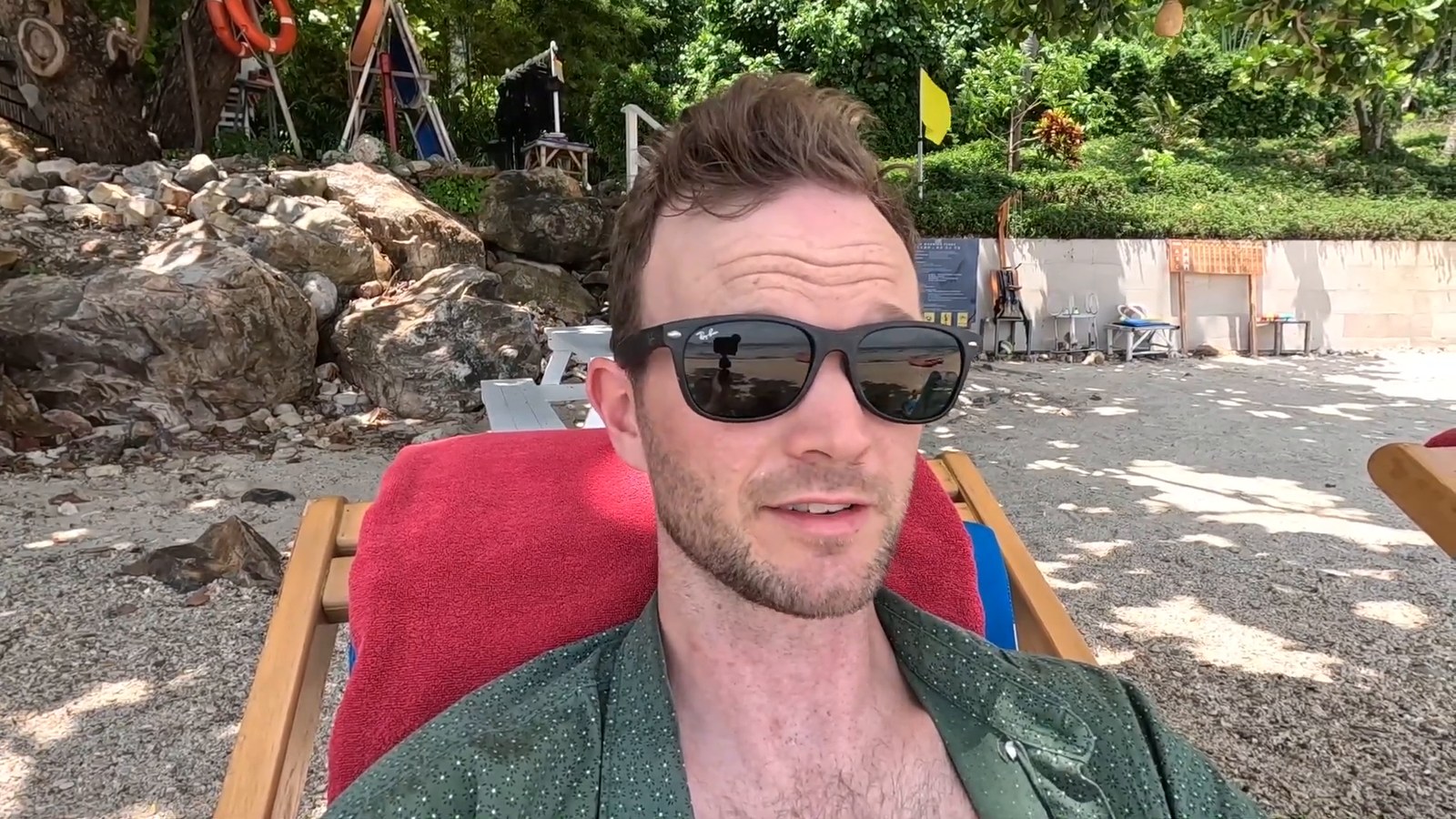 A man in sunglasses lying on a red-towel sun lounger on a pebbly beach with rocks and green trees behind.