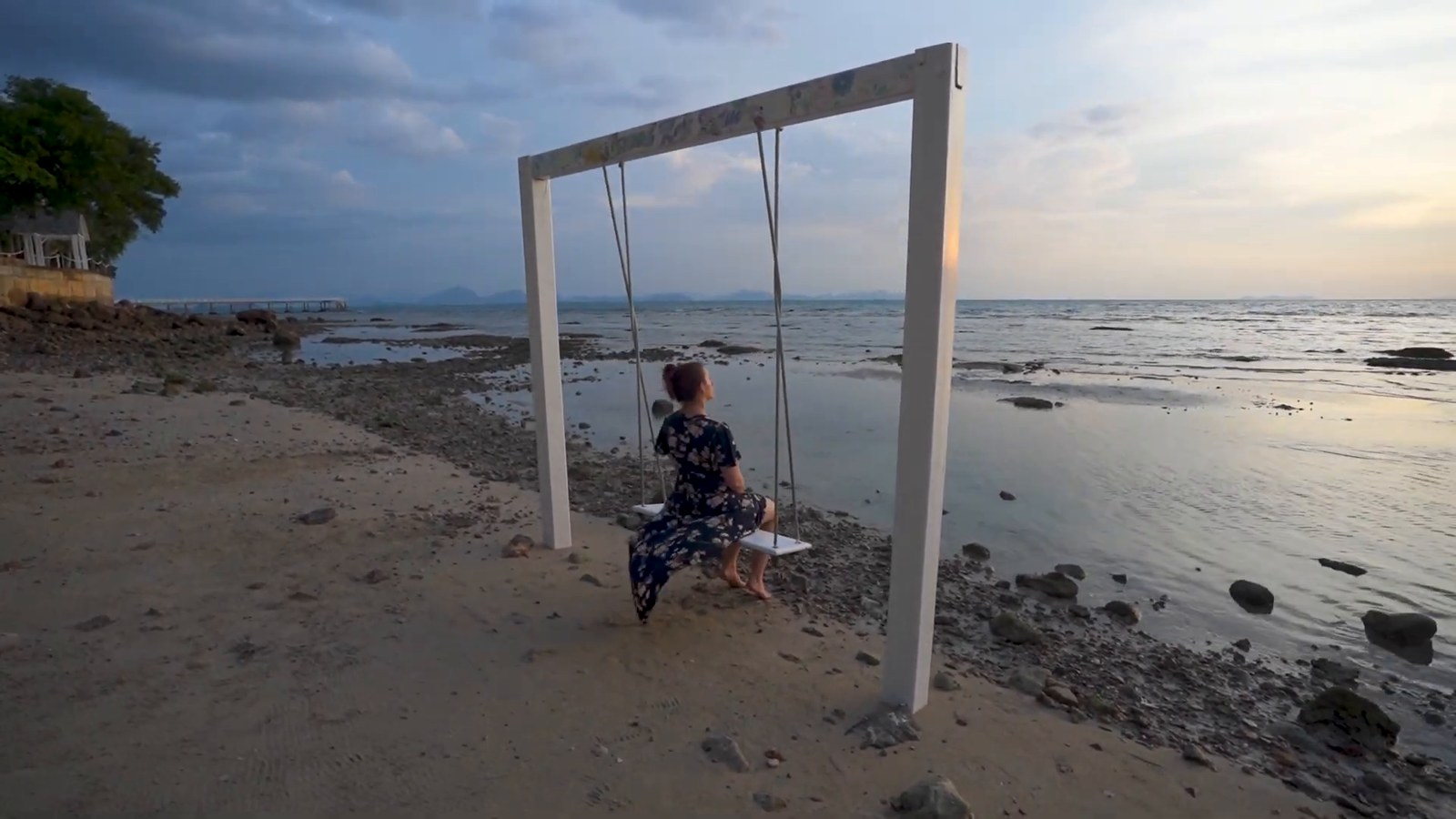A woman sitting on a swing hung from a white frame on a rocky beach at dusk, looking out over calm water.
