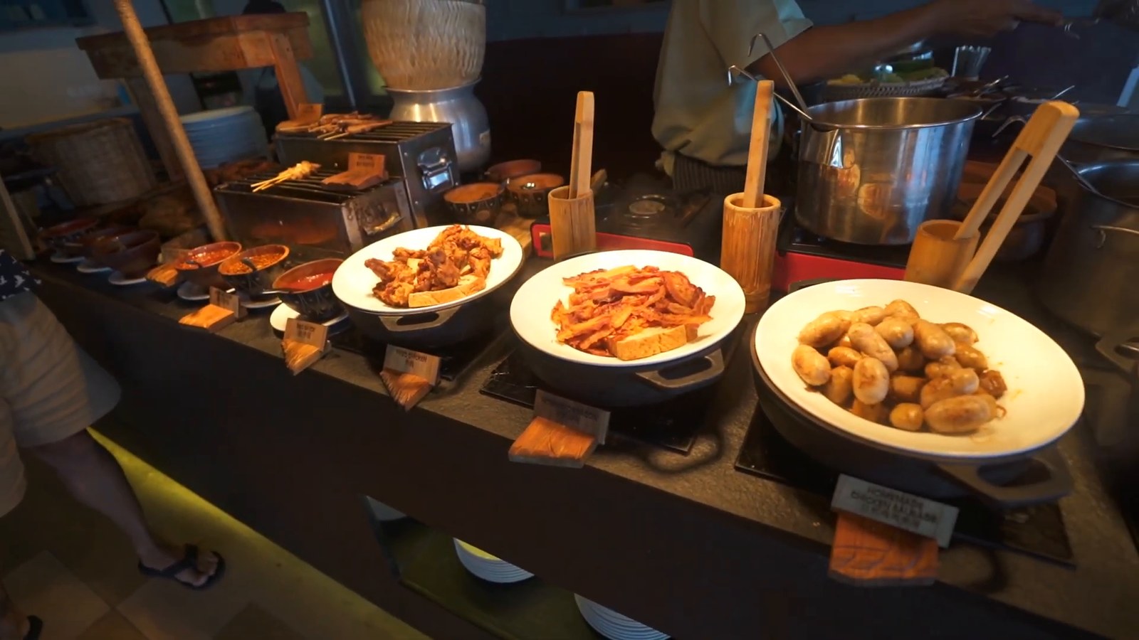 A breakfast buffet counter with cast-iron pans of fried food, sausages and stir-fried dishes with small wooden labels.