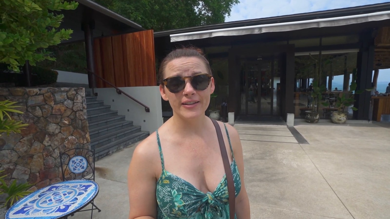 A woman in sunglasses and a green patterned top standing outside a wood-and-glass restaurant building with a stone wall and steps.