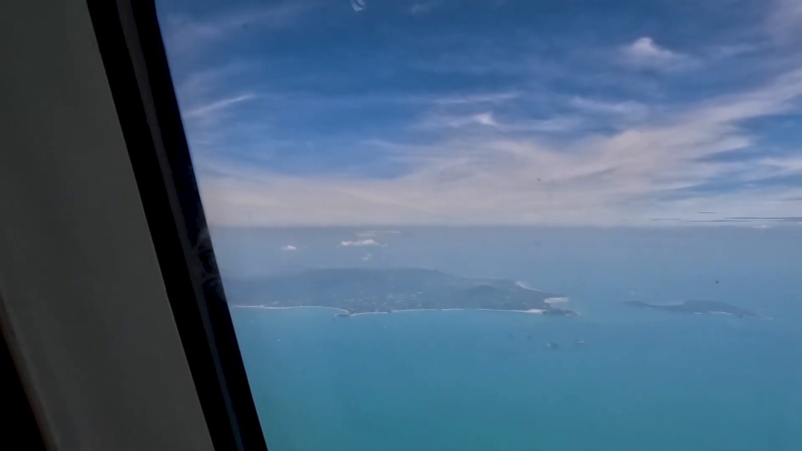 A view from an airplane window over blue sea and a coastline with small islands.