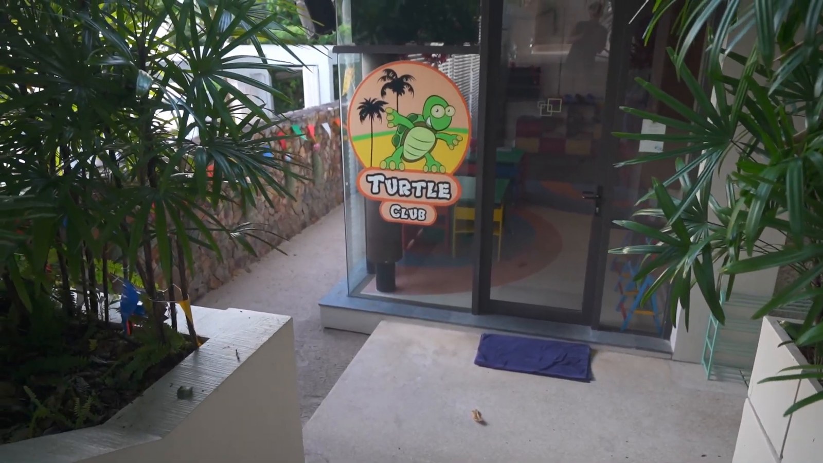 The glass entrance to the Turtle Club kids club, with a smiling turtle logo, palms and a stone wall.
