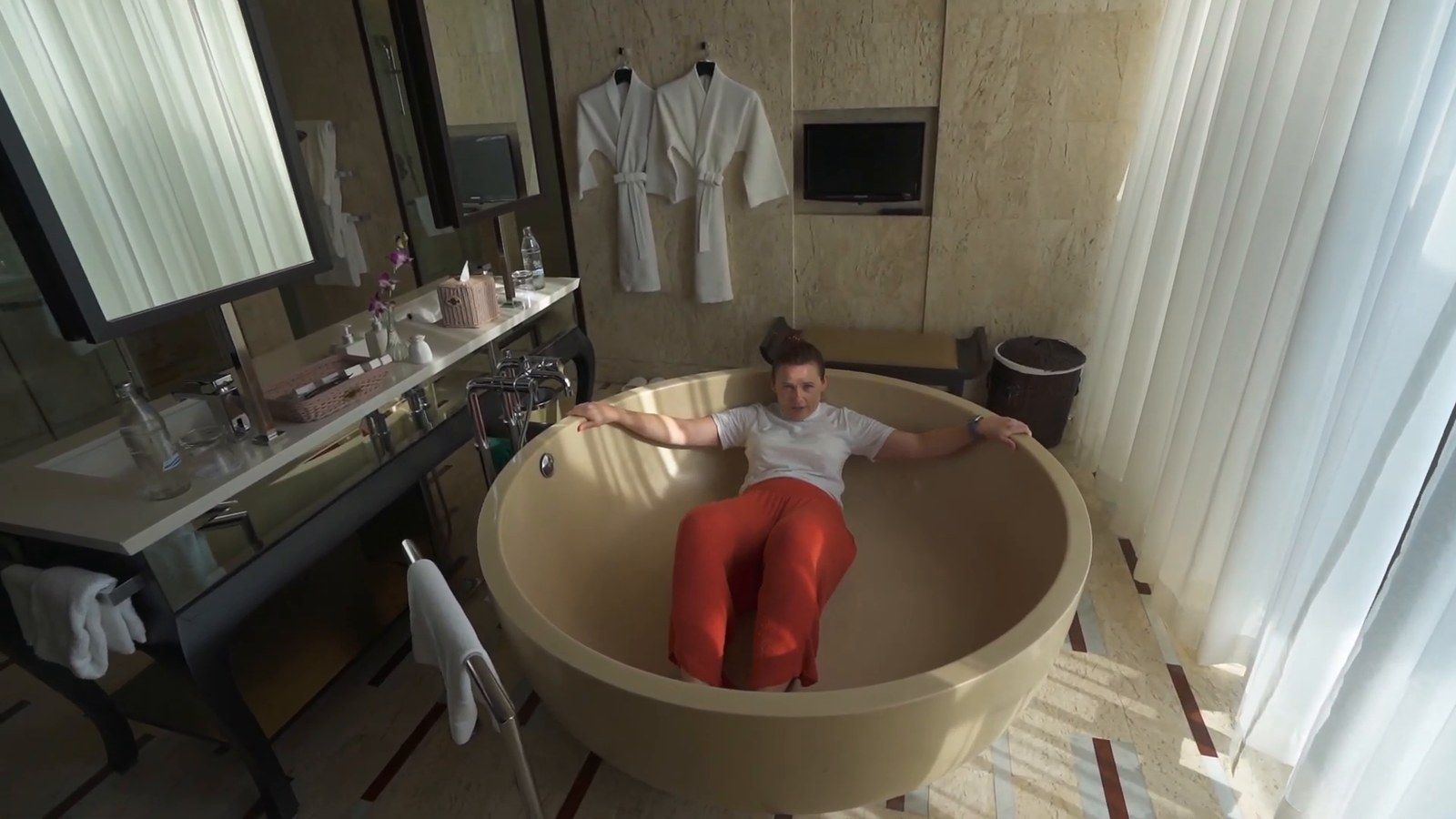 A woman lying back in a large round bathtub in a bathroom with pale stone walls, with a double vanity and robes hanging on the wall.