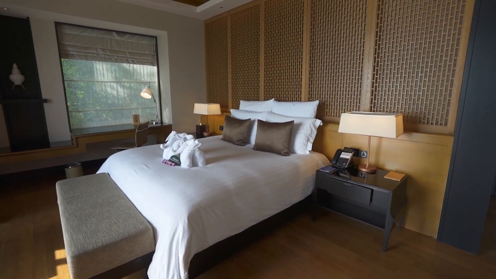 A villa bedroom with a king bed, wooden lattice headboard wall, bedside lamps, a bench at the foot of the bed and a folded towel on the bed.