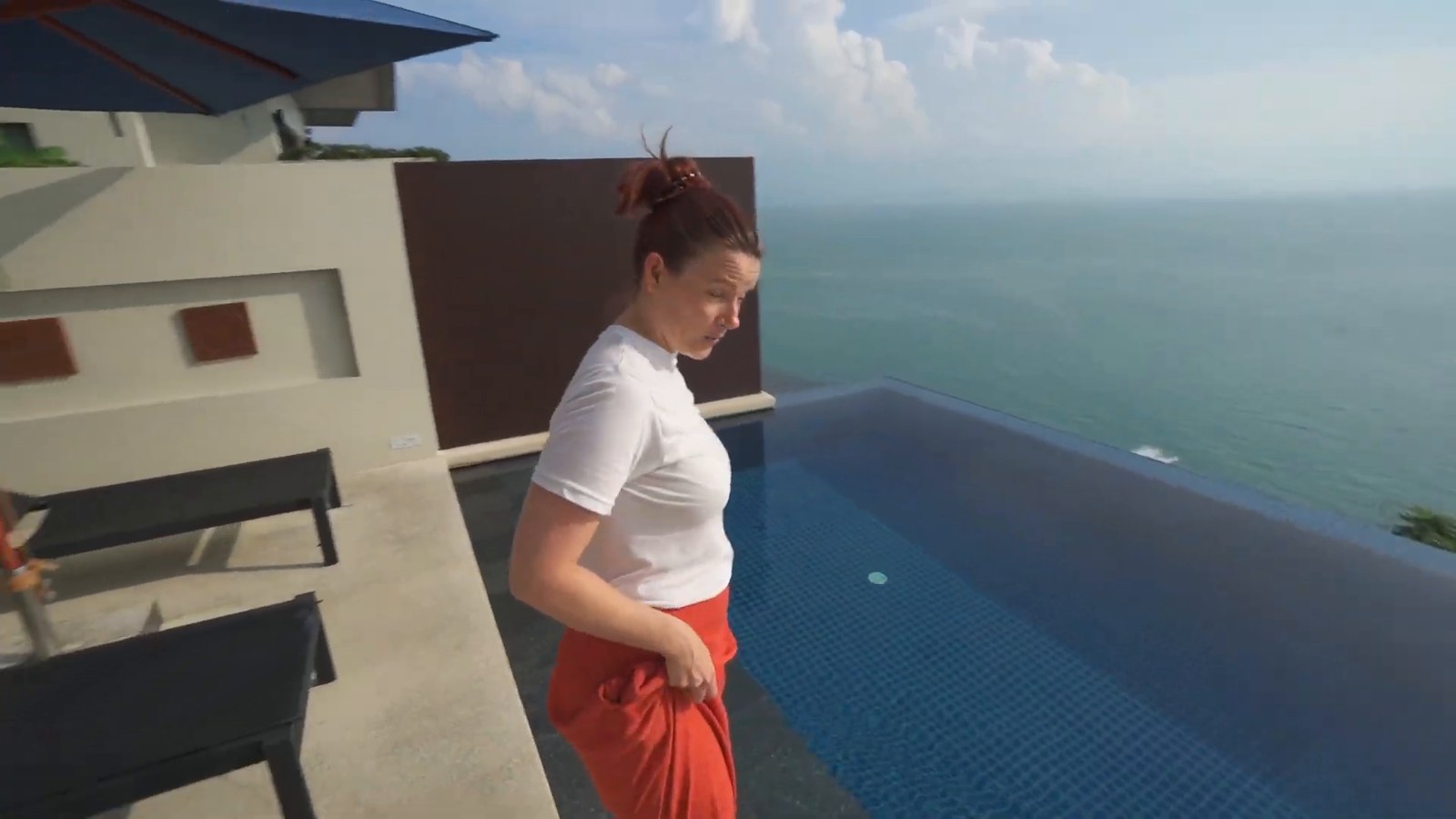 A woman standing beside a blue-tiled private infinity pool with loungers and a view over the sea.