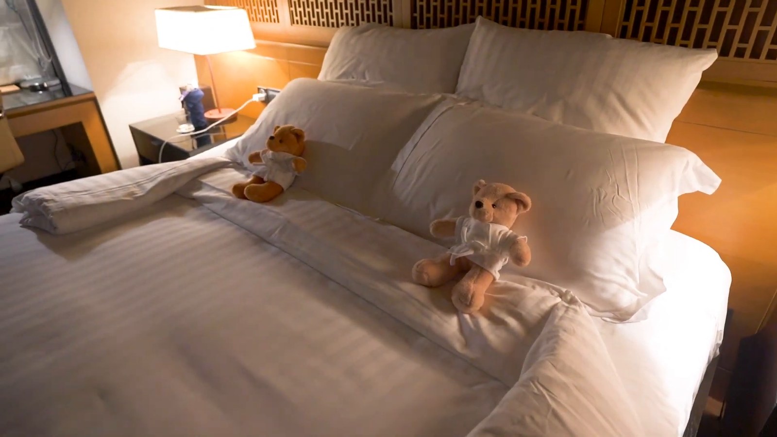 Two small teddy bears in white shirts sitting on a white-sheeted bed with pillows and a lit bedside lamp.