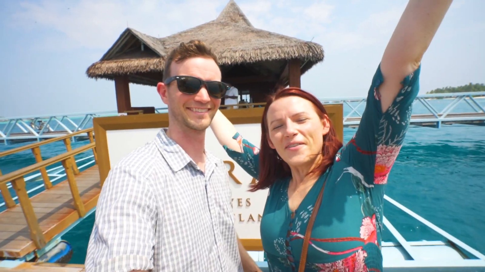 Two travelers smiling on a wooden dock in front of the resort sign, with a thatched hut and turquoise water behind them.