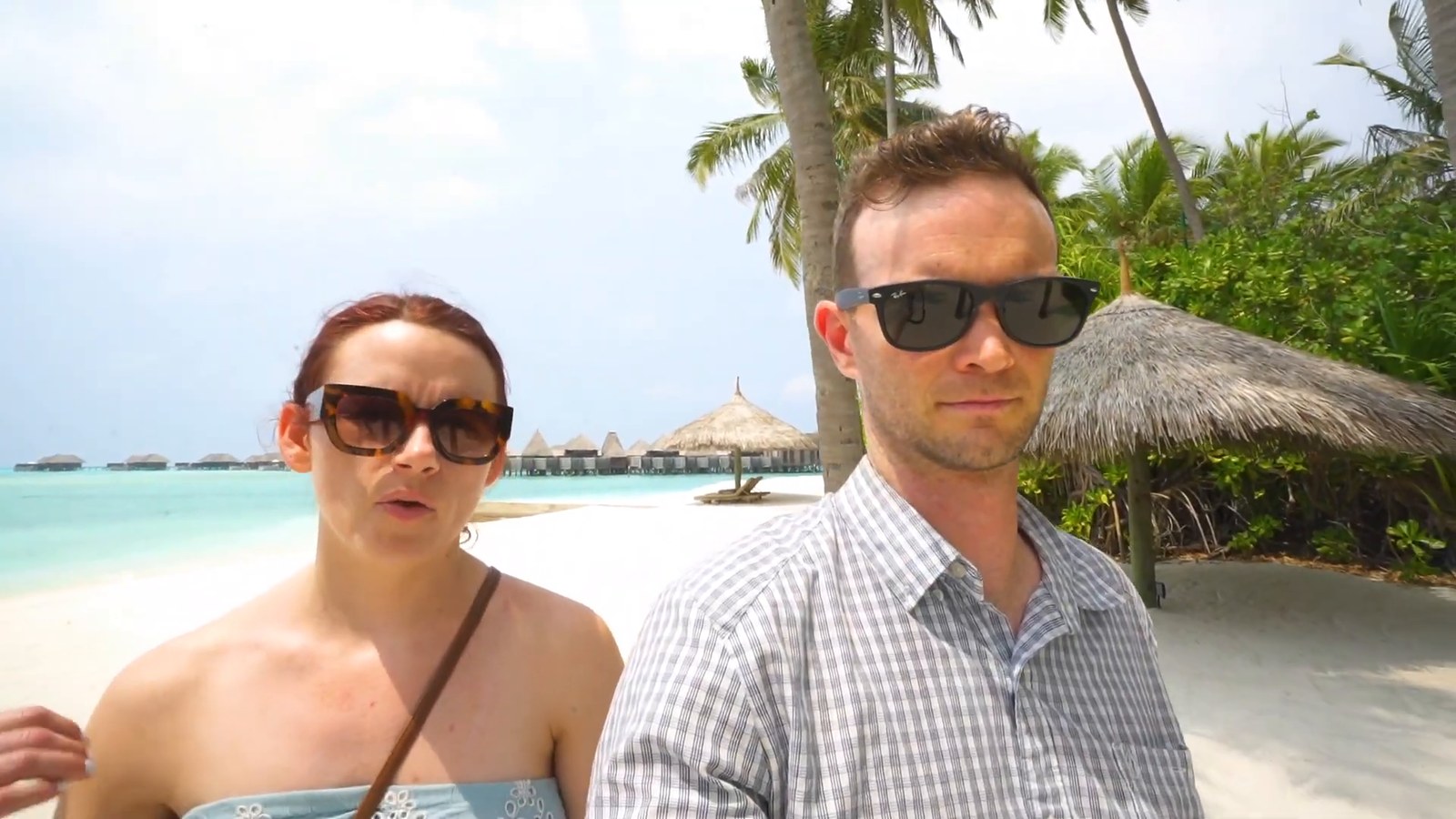 Two travelers on a white-sand beach with overwater villas and palm trees behind them.