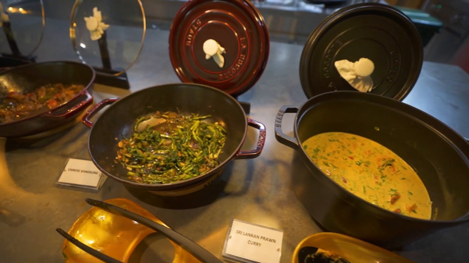 Cast iron pots of curry and stir-fried greens with small labels on a buffet counter.