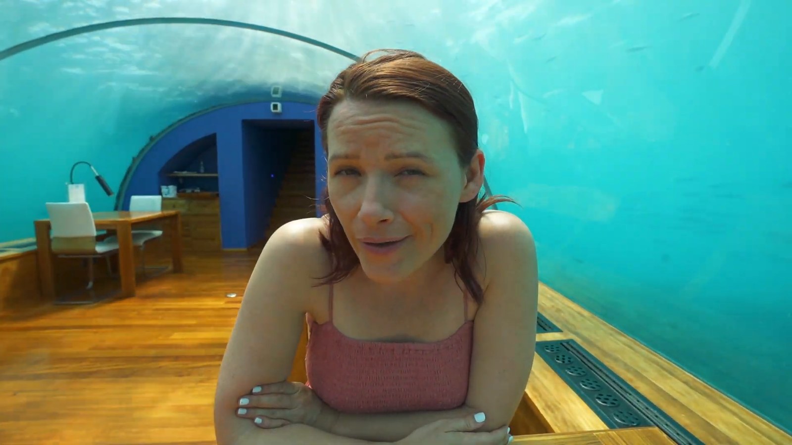 A woman smiling inside a glass-walled underwater restaurant, with wooden floors, a table and blue-green water behind her.