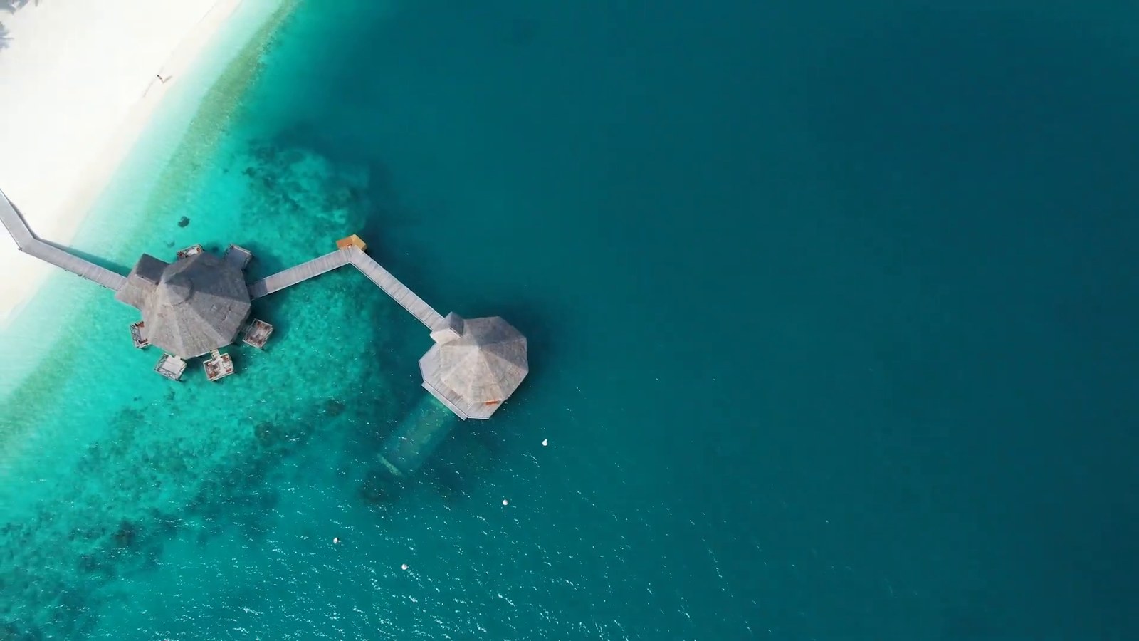Aerial view of two thatched overwater buildings joined by a wooden walkway over turquoise water.