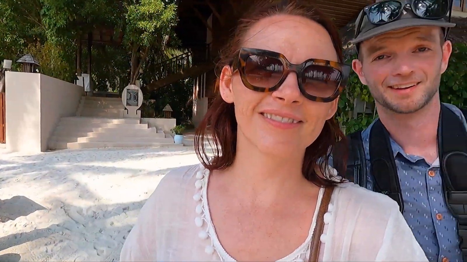 Two travelers smiling on a white-sand path in front of stone steps and tropical trees.