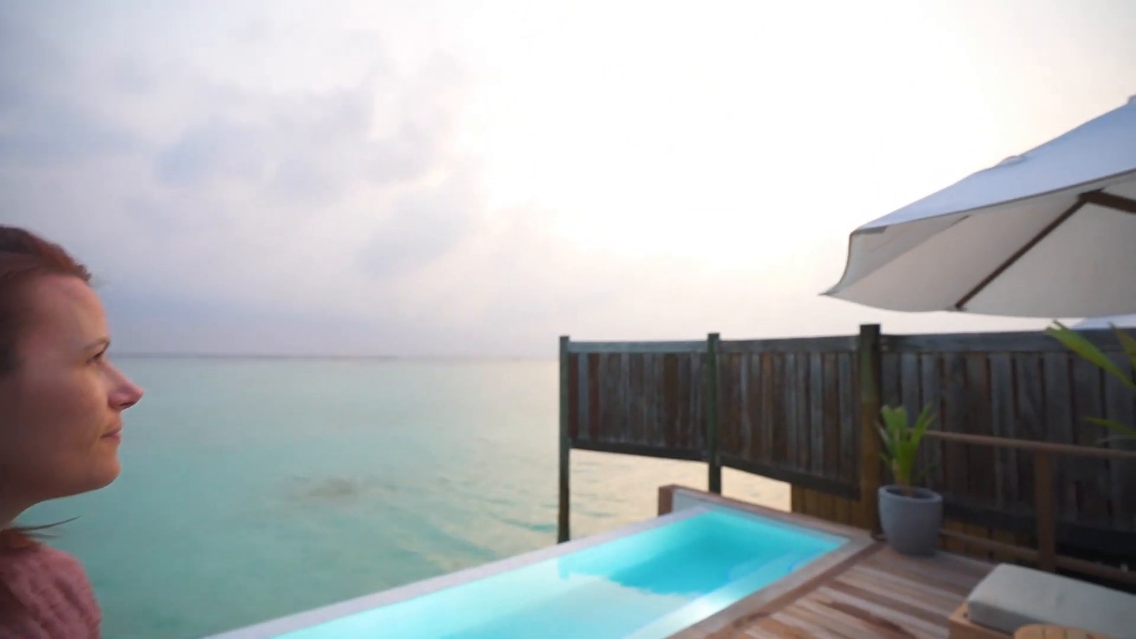A woman looking out at a hazy sunset over the water from a villa deck with an infinity-edge pool and a sun umbrella.