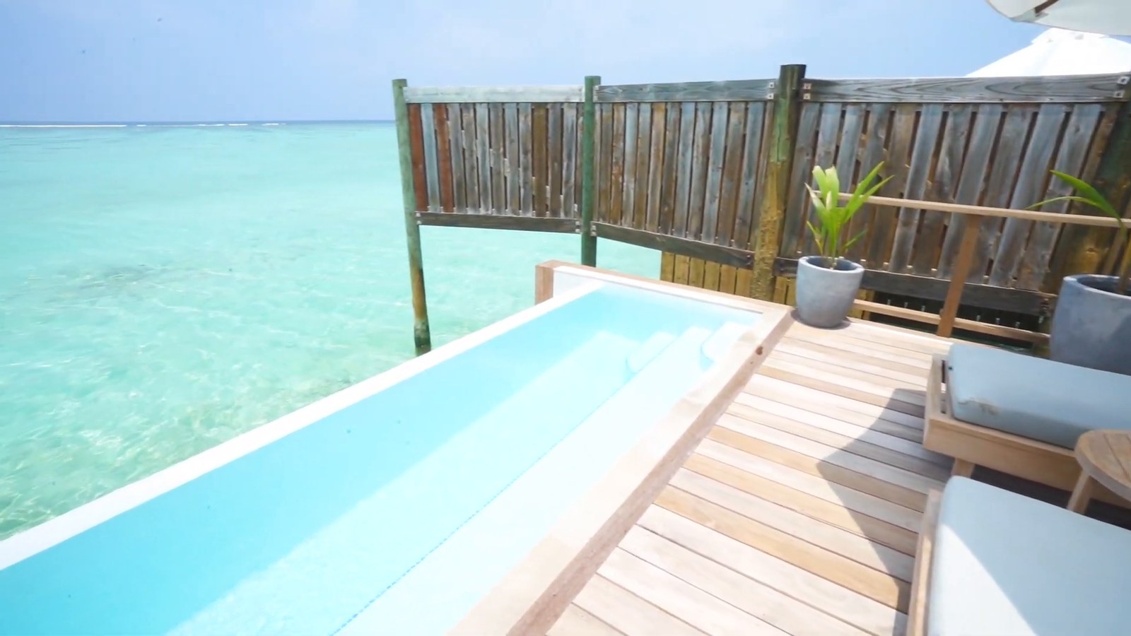 A water villa deck with an infinity-edge pool, loungers and a wooden railing over turquoise water.