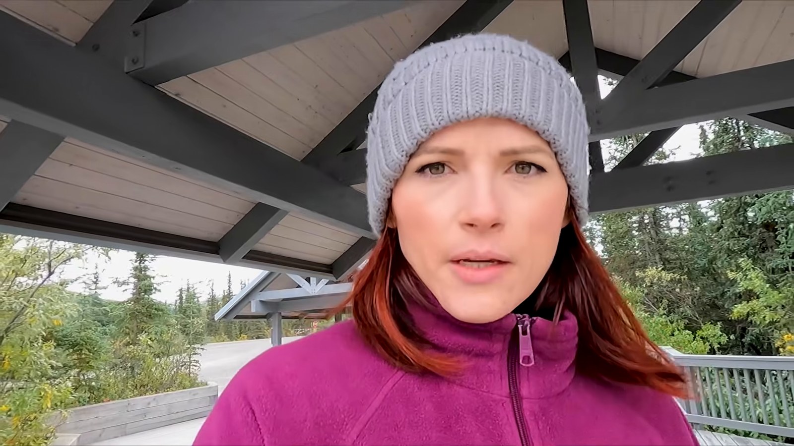 A woman in a grey knit beanie and a fleece standing under a covered shelter at a rest stop, with trees behind her.