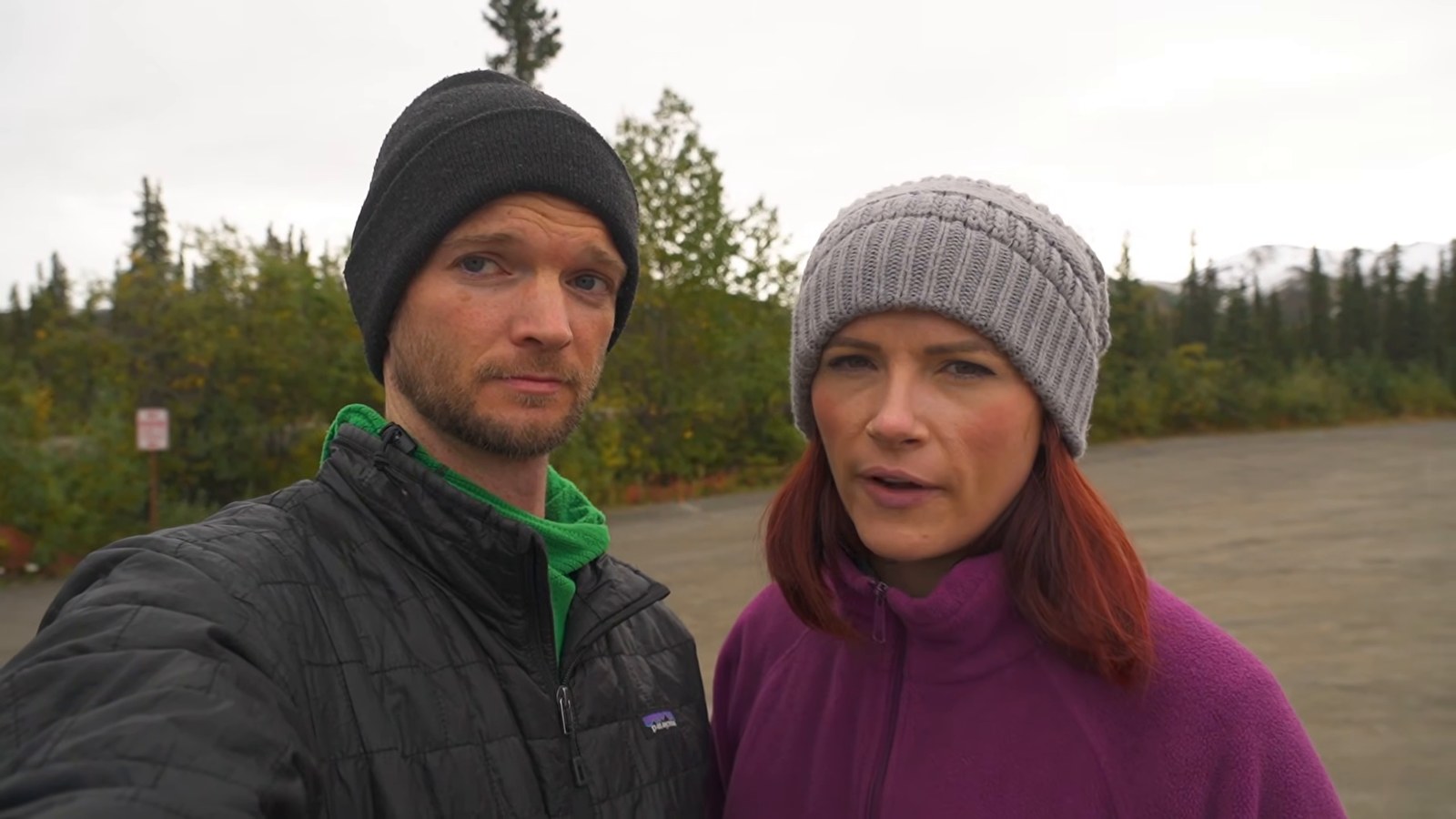 A man in a black beanie and a woman in a grey beanie standing in a gravel lot surrounded by spruce trees on a cloudy day.