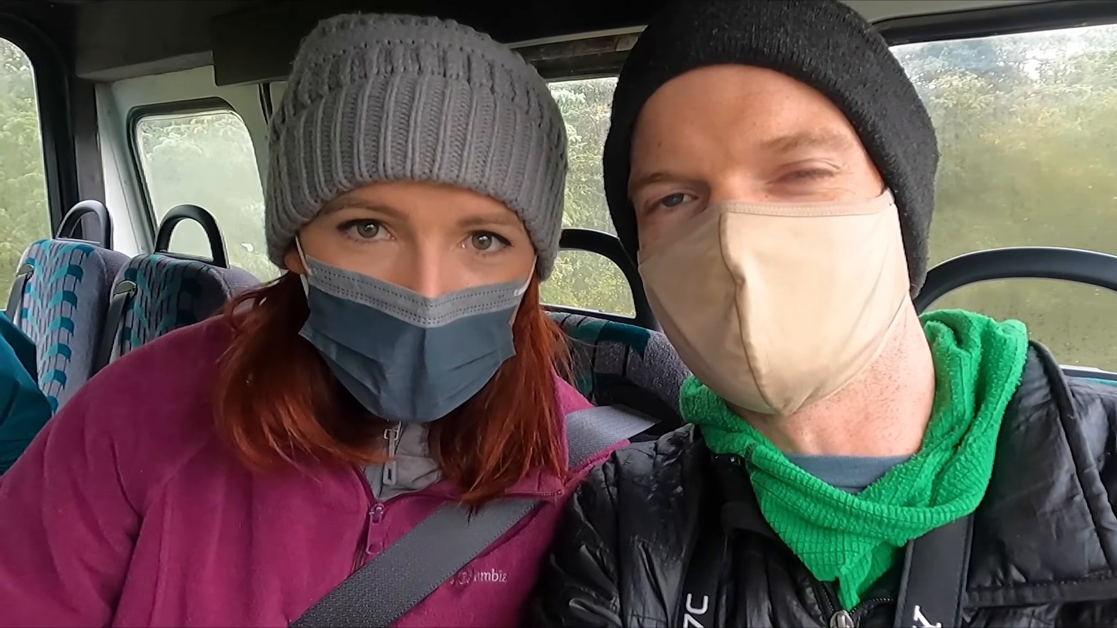 A man and a woman in beanies and face masks taking a selfie in their seats on a Denali tour bus.
