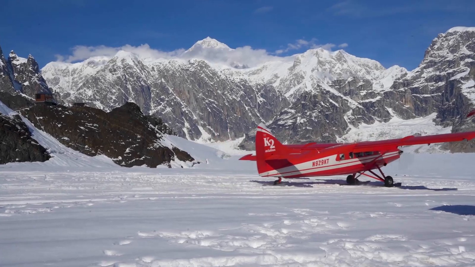 A red ski plane parked on a snowfield surrounded by rocky, snow-covered peaks.