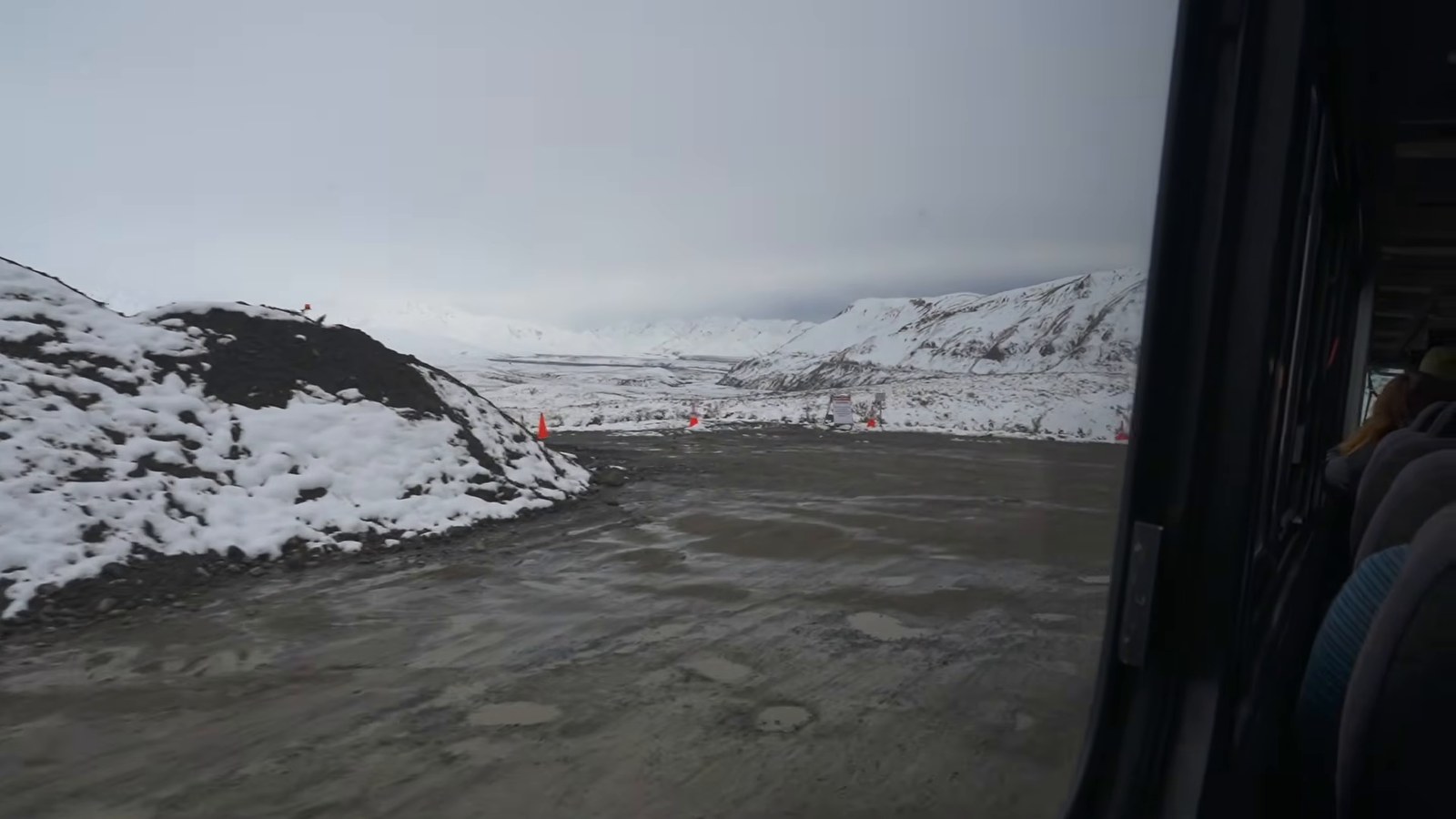Snow-covered hills and a gravel road seen through a bus window under grey skies.