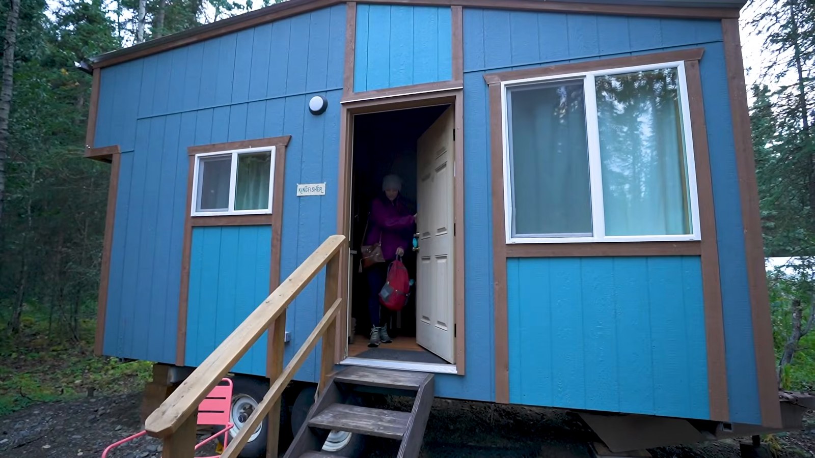 A small blue tiny house on a trailer with wooden steps, surrounded by spruce trees.