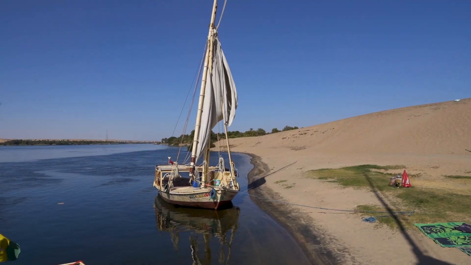 A white felucca sailboat moored beside a sand dune on the bank of the Nile under a clear blue sky.