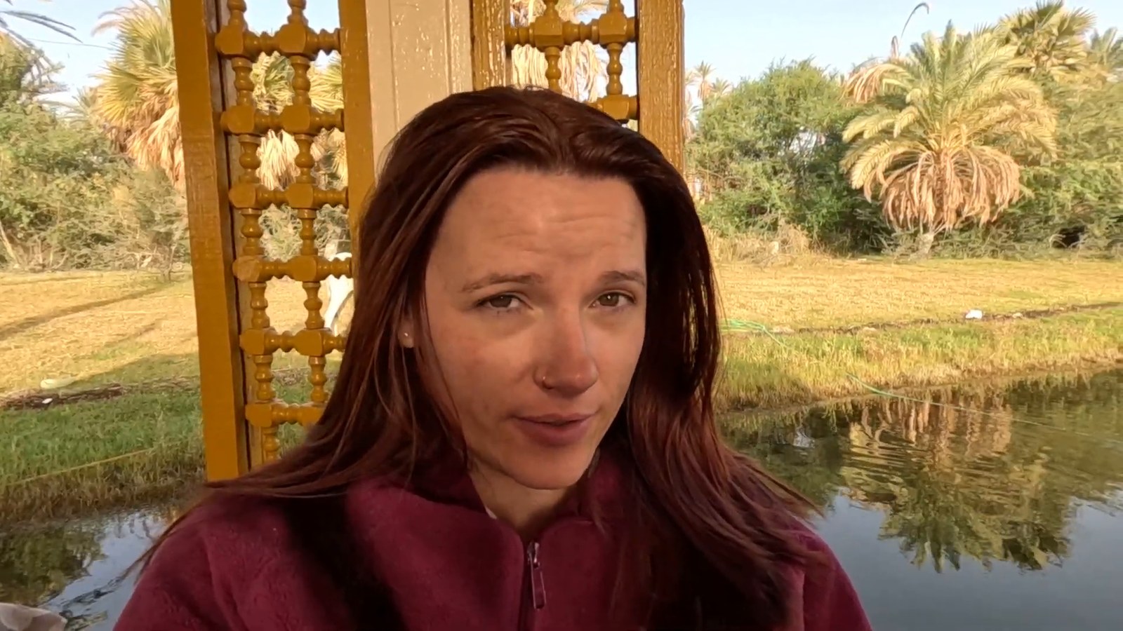 A woman in a red fleece on the deck of a felucca in the early morning, with palm trees reflected in the Nile behind her.