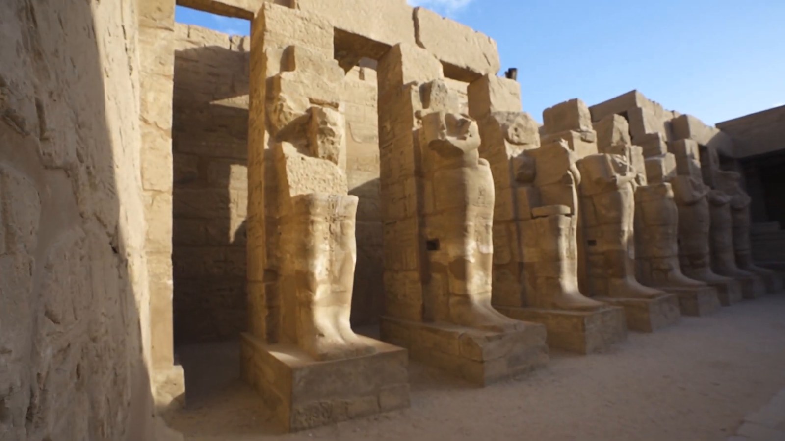 A row of tall carved stone statues standing in niches along a sunlit temple court at Karnak.