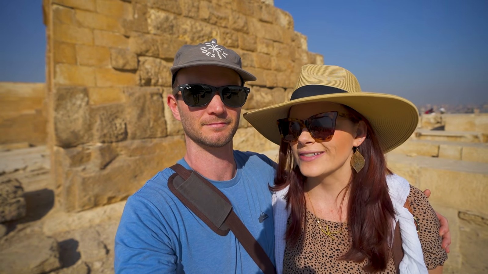 A man in a cap and sunglasses and a woman in a straw hat smiling beside huge ancient stone blocks at Giza.