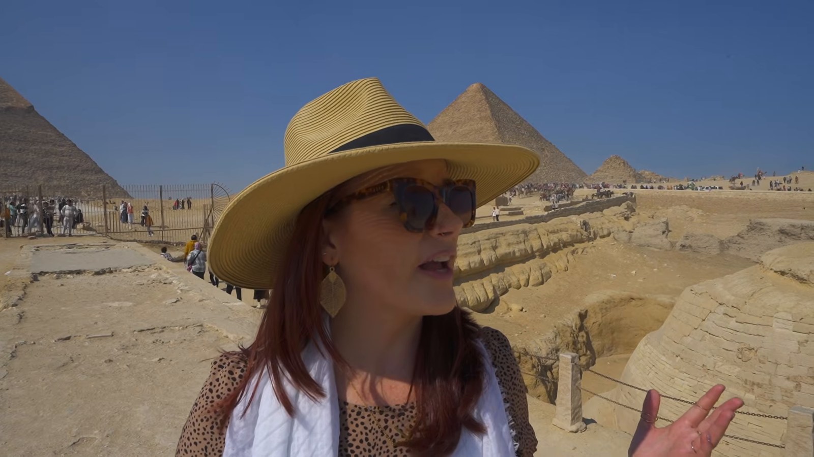 A woman in a wide straw hat and sunglasses standing on the Giza plateau with two of the great pyramids behind her under a blue sky.