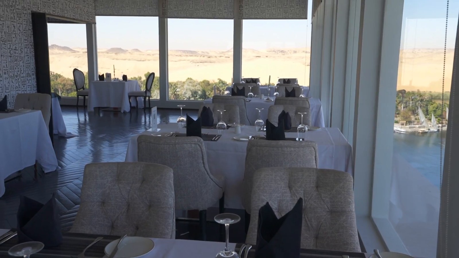 An empty restaurant with white tablecloths and floor-to-ceiling windows looking out over the Nile and the desert at Aswan.