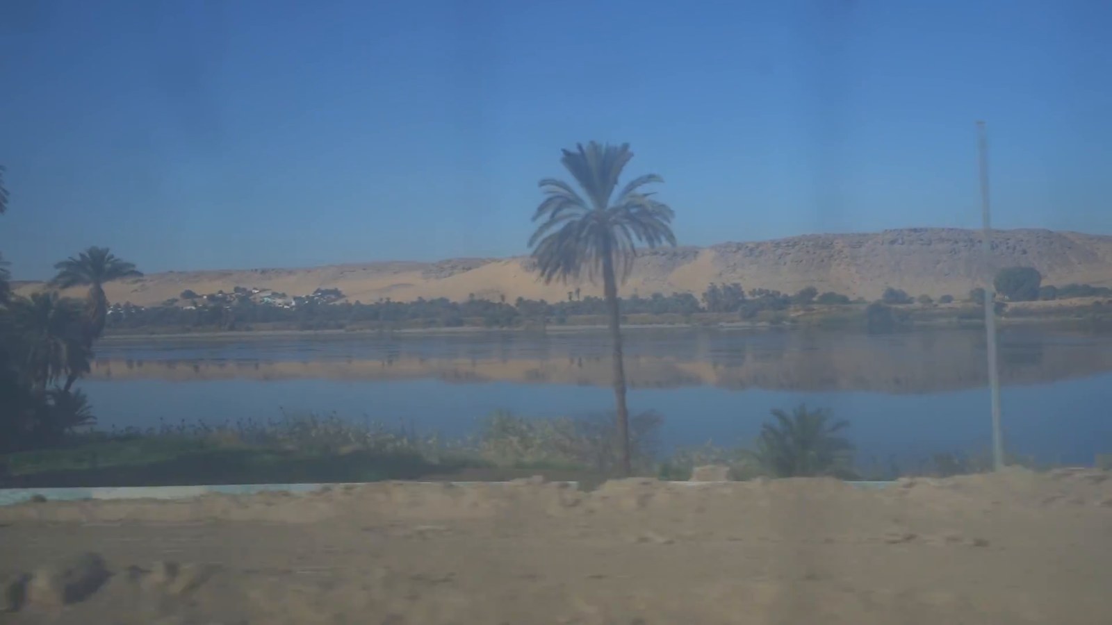 The Nile seen from a train window in the morning: still water, a palm tree and sand dunes on the far bank.