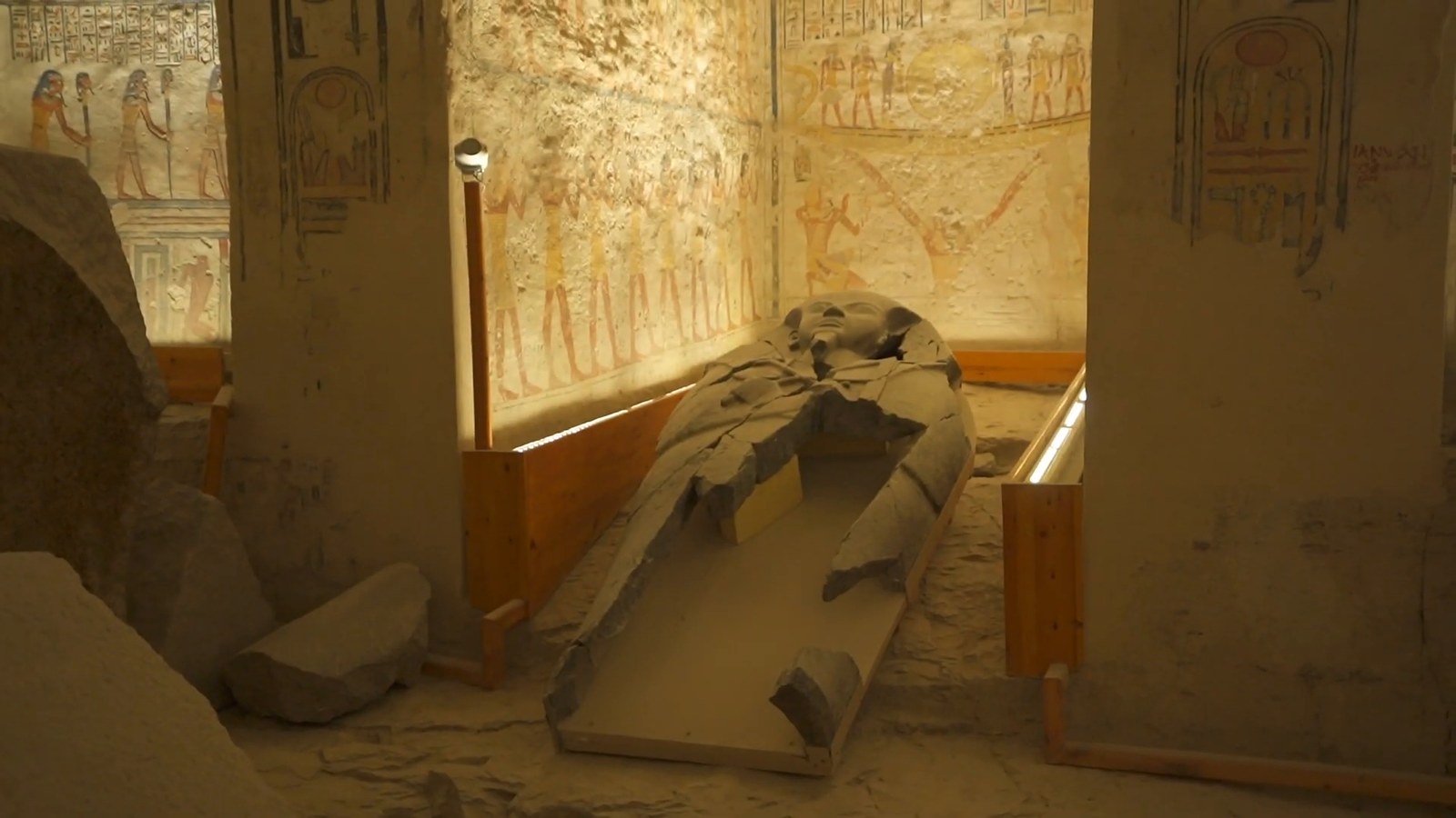 A broken stone sarcophagus lid lying in a burial chamber with painted walls, behind a wooden barrier.