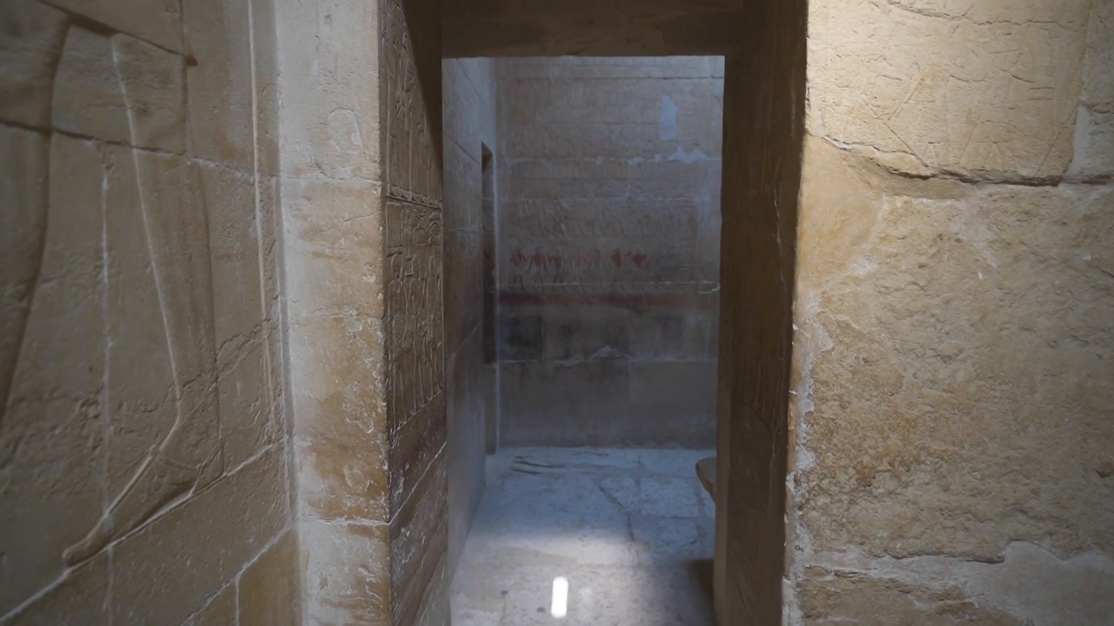 A narrow doorway between stone walls carved with reliefs, leading into a chamber with painted hieroglyphs in a Saqqara tomb.