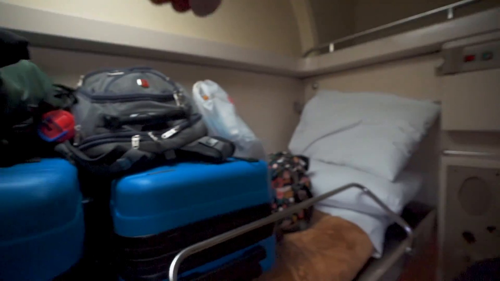 A small sleeper-train cabin with a made-up berth, white pillow and suitcases and backpacks piled on the seats.