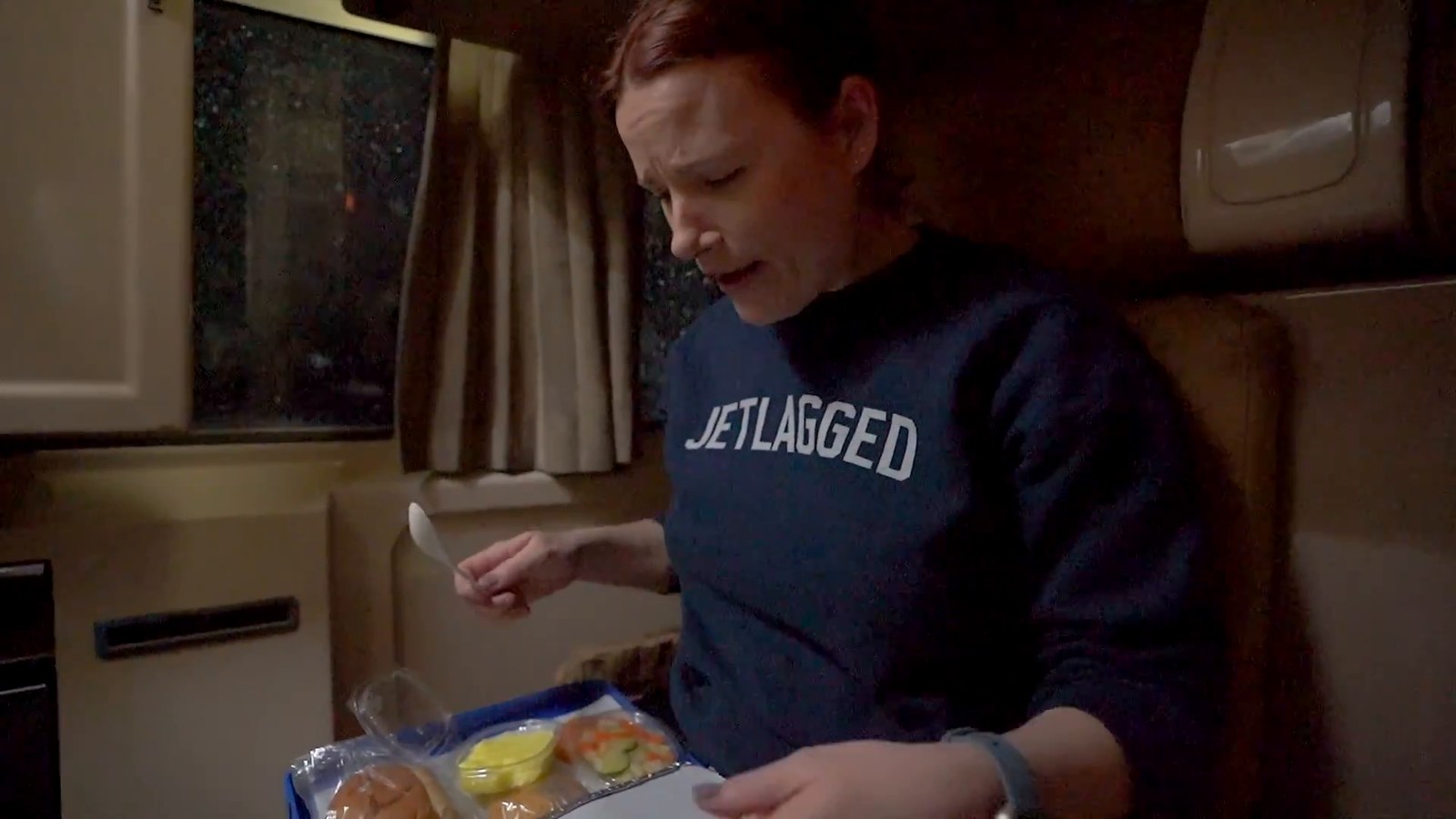 A woman in a navy sweatshirt looking down at a dinner tray with a roll, rice and a yellow pudding in a train cabin.