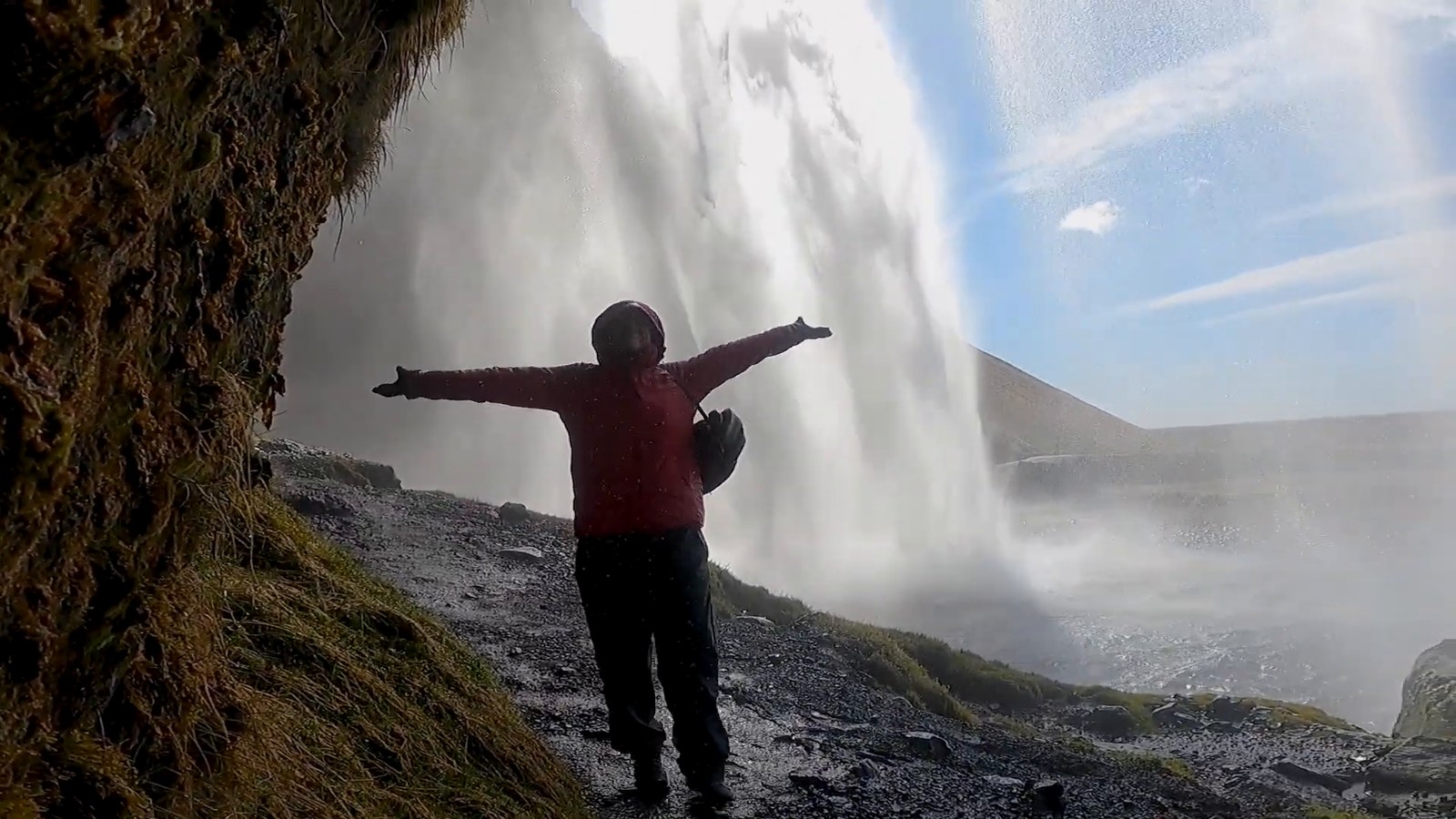 A person in a red jacket with arms outstretched on the path behind Seljalandsfoss waterfall.