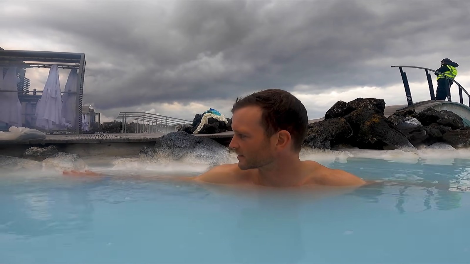 A man relaxing in the milky blue water of the Blue Lagoon under a cloudy sky, with dark lava rock behind him.