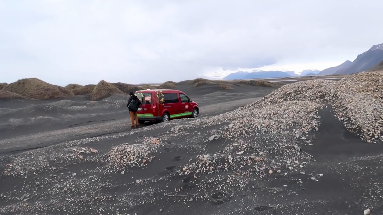 A red campervan parked on black sand dunes with mountains and cloud in the distance.