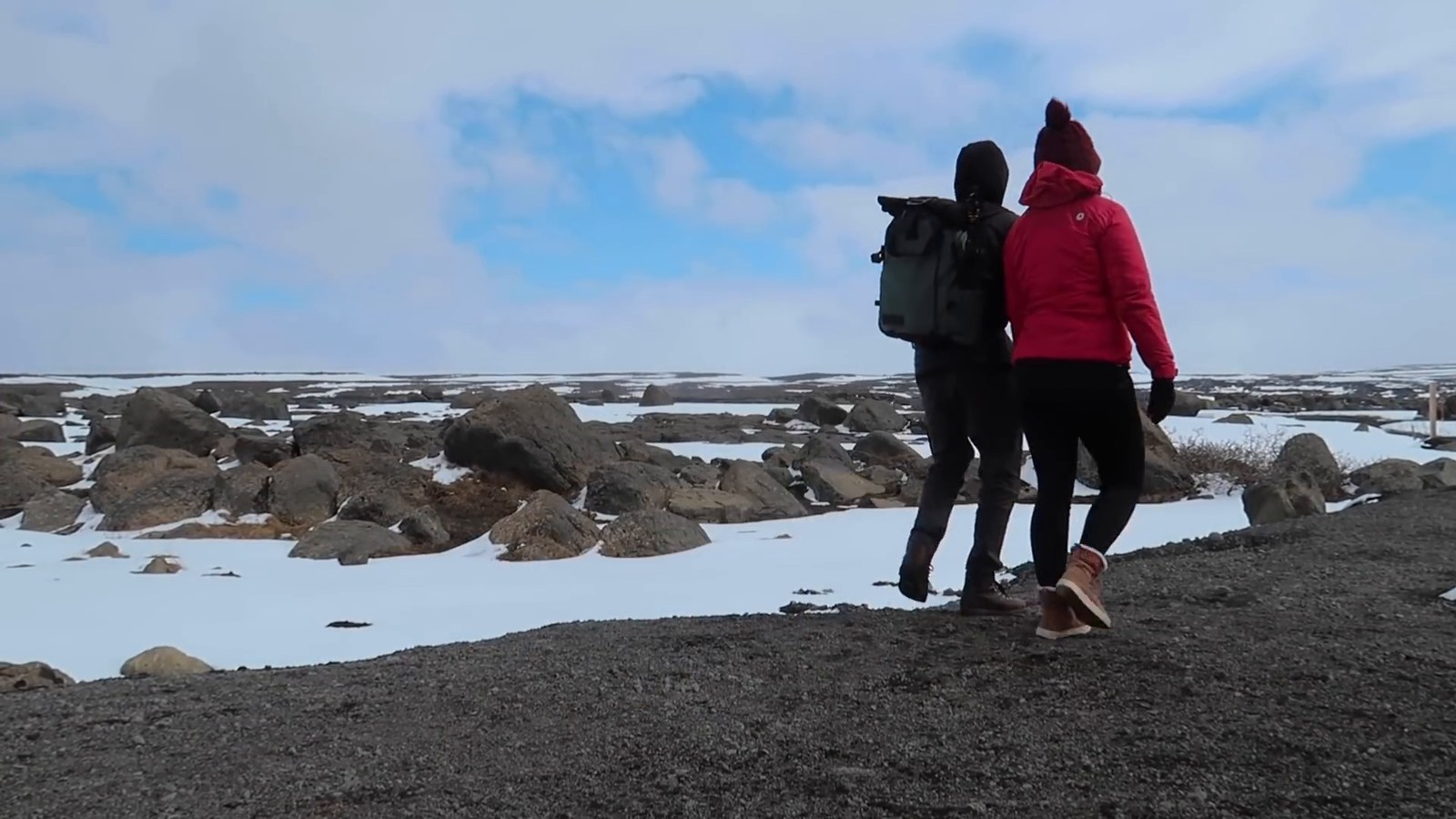 Two hikers in winter jackets walking along a rocky rim with patches of snow towards the Dettifoss viewpoint.