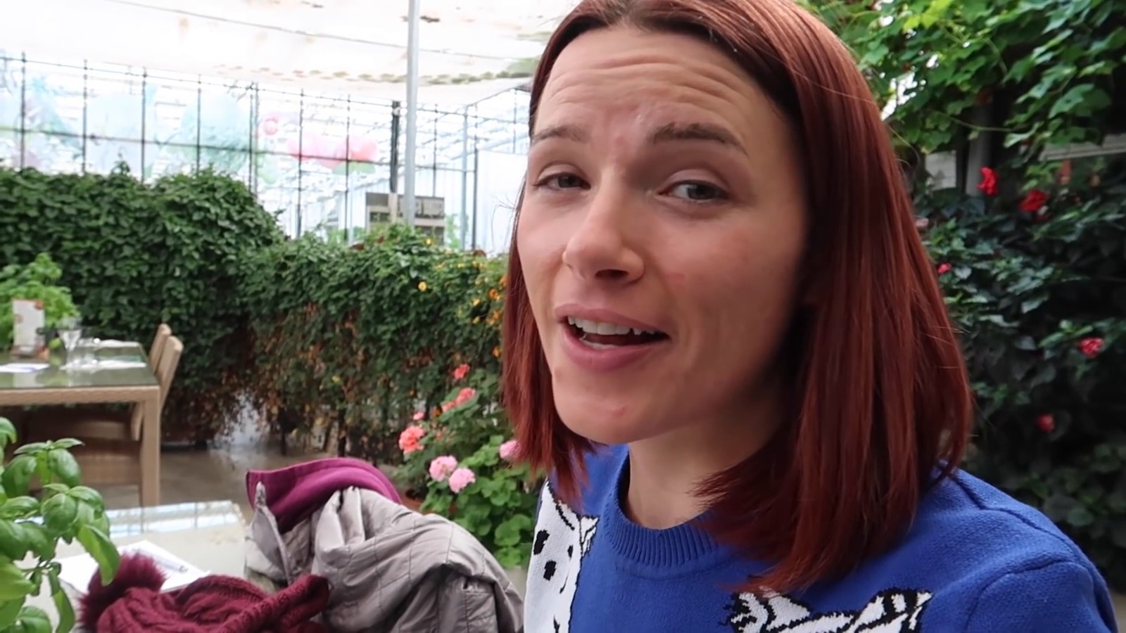 A woman smiling at the camera inside a bright greenhouse restaurant full of plants, with a table and chairs behind her.