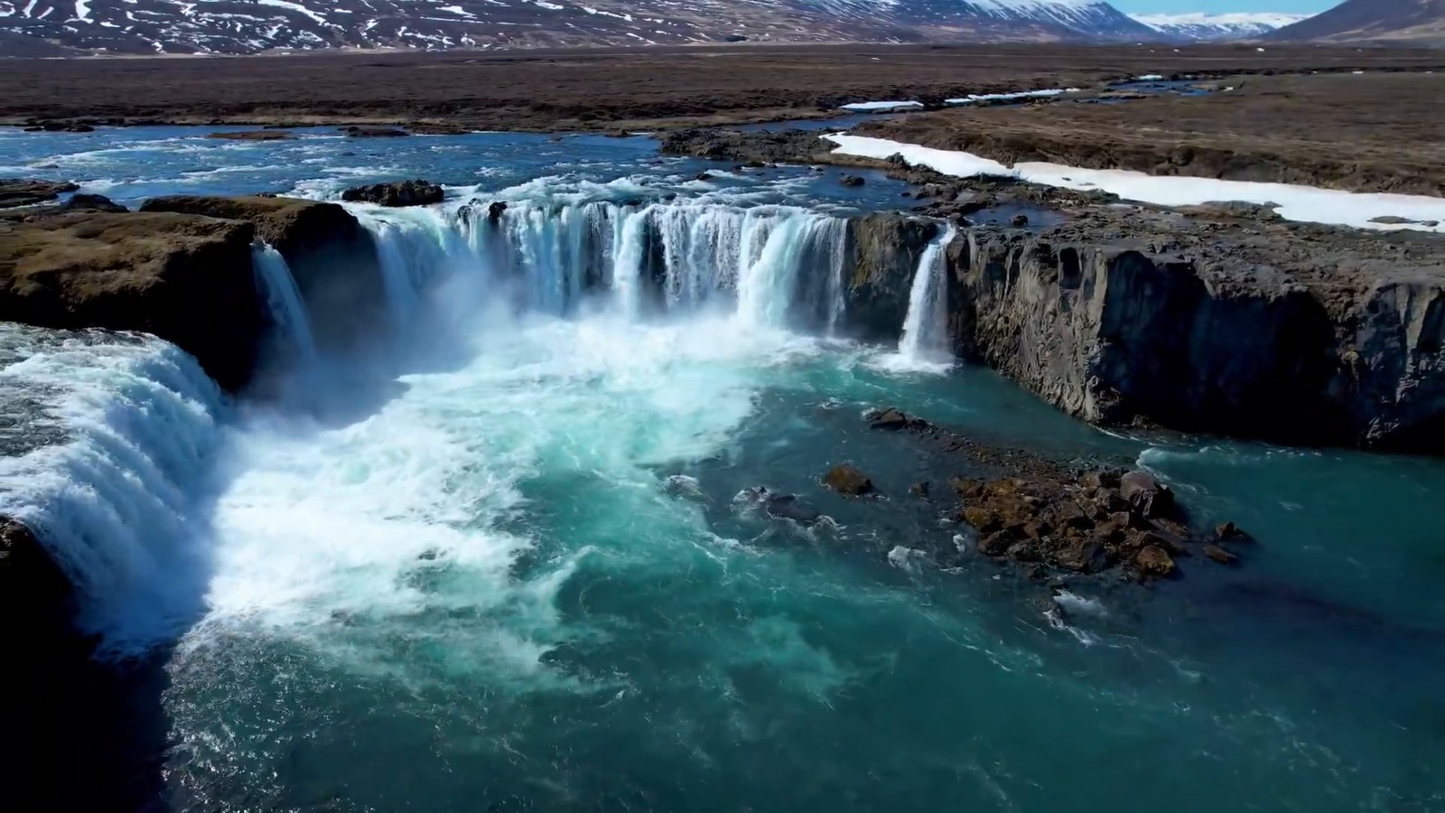 Aerial view of the horseshoe-shaped Goðafoss waterfall with turquoise water below the falls.
