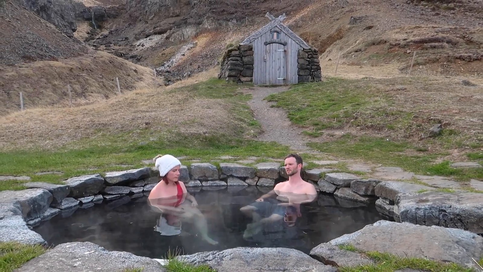 Two people soaking in a round stone-lined hot spring with a small wooden hut and a grassy hillside behind them.