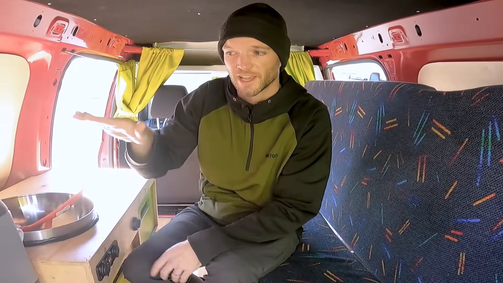Kevin sitting on the bench seat inside the small red campervan, with the kitchen unit and orange walls behind him.