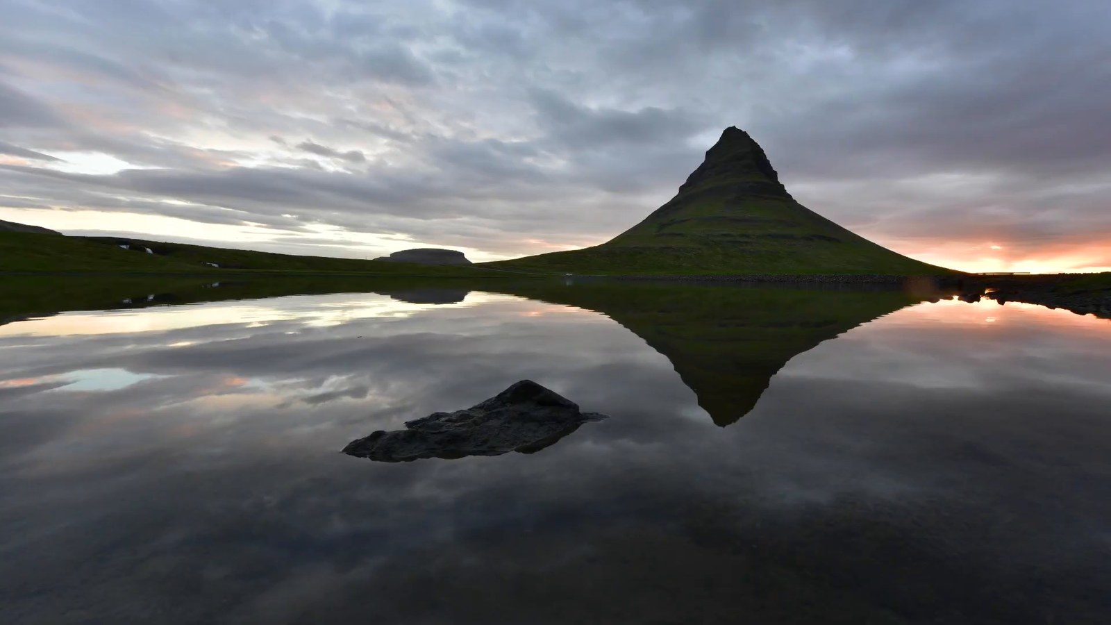 Kirkjufell mountain reflected in still water under a cloudy sunset sky.