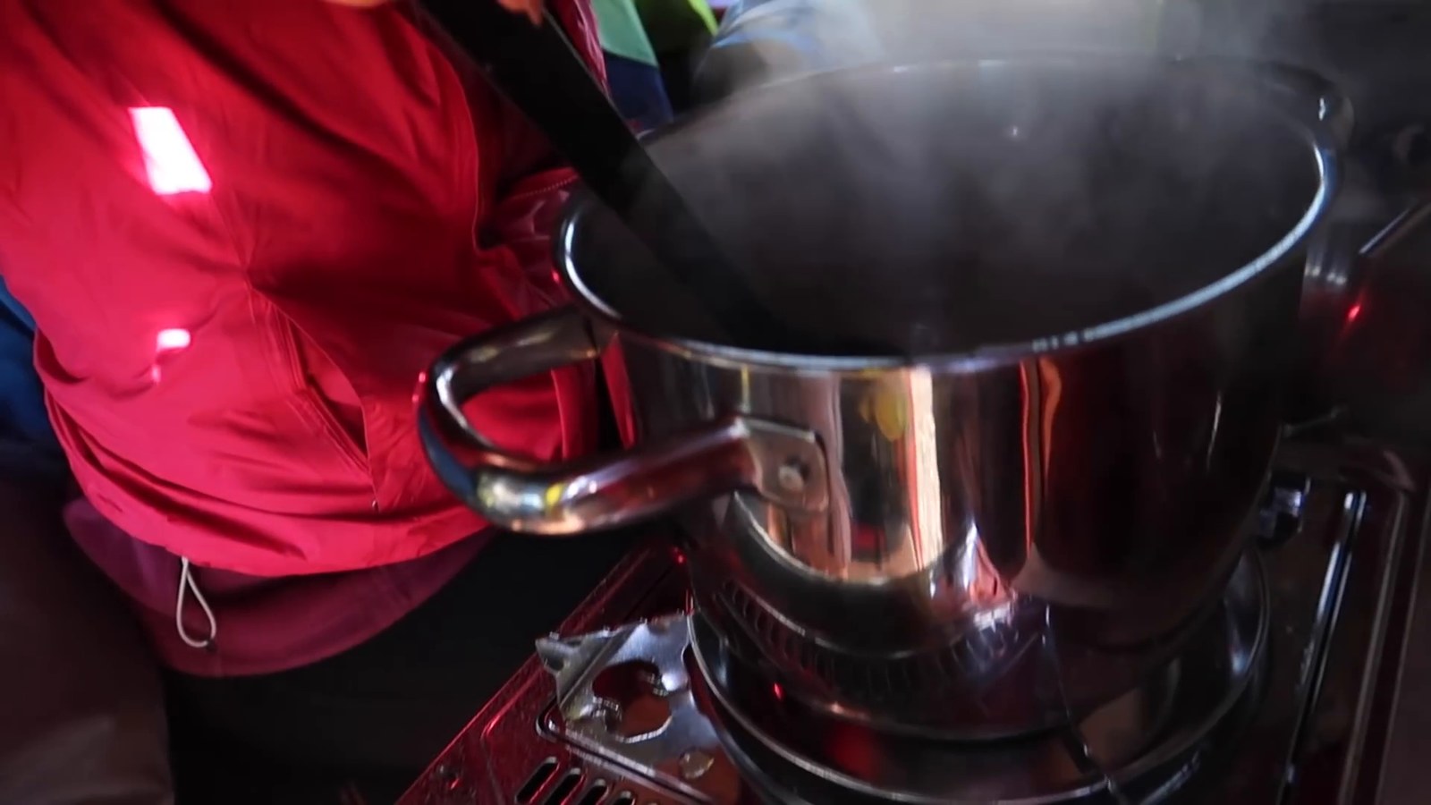 A steaming pot on a small one-burner camping stove next to a person in a red jacket.
